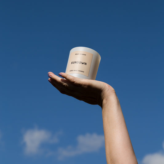Hand holding a 'Sundown' jar candle against a blue sky