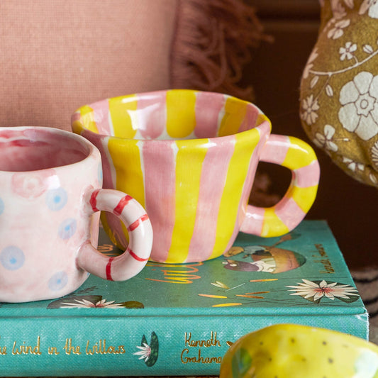 The Bloomingville Nini Cup in Pink and Yellow Stripe displayed on a book alongside another pink cup with a pink tufted cushion in the background.