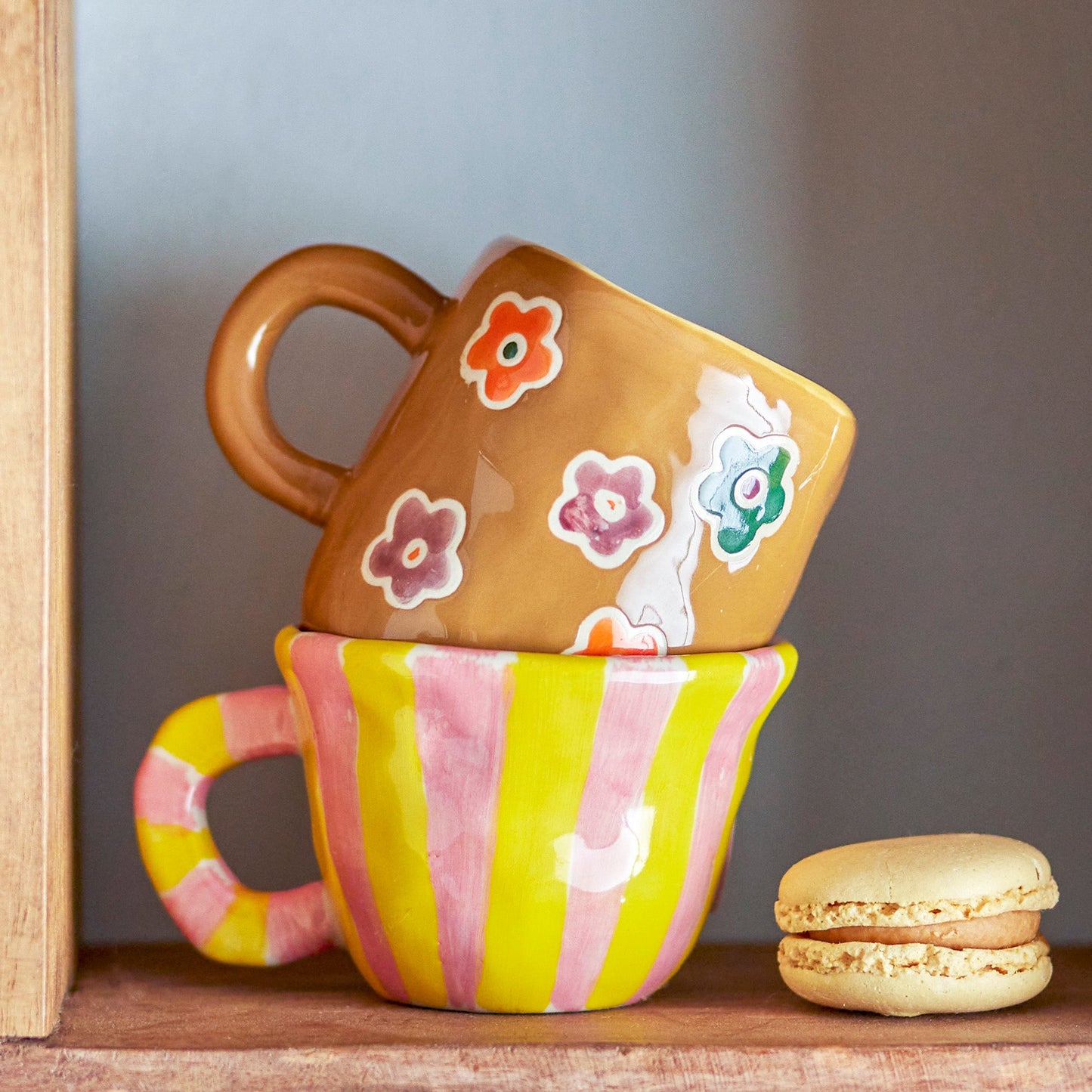 Bloomingville Nini Cup in Pink and Yellow Stripe sat on a shelf with another Bloomingville cup with flower detail sat inside it with a biscuit sat on shelf next to them.