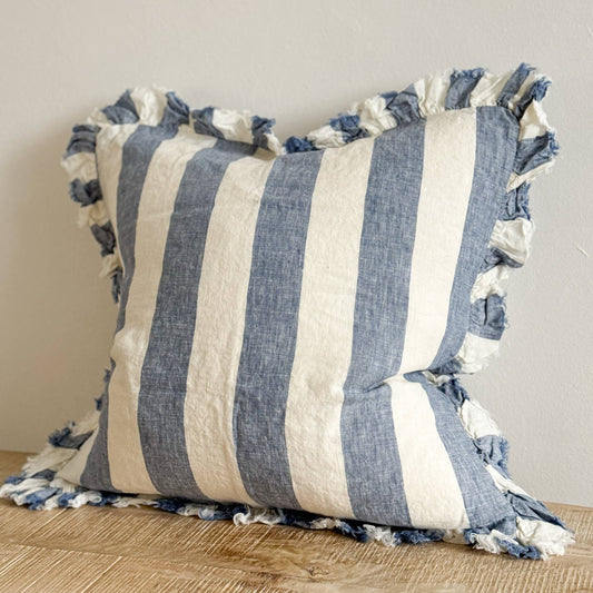 Ellenvale Hallie Ruffled Edged Linen Cushion in Blue Wide Stripe displayed on a bench against a white wall.