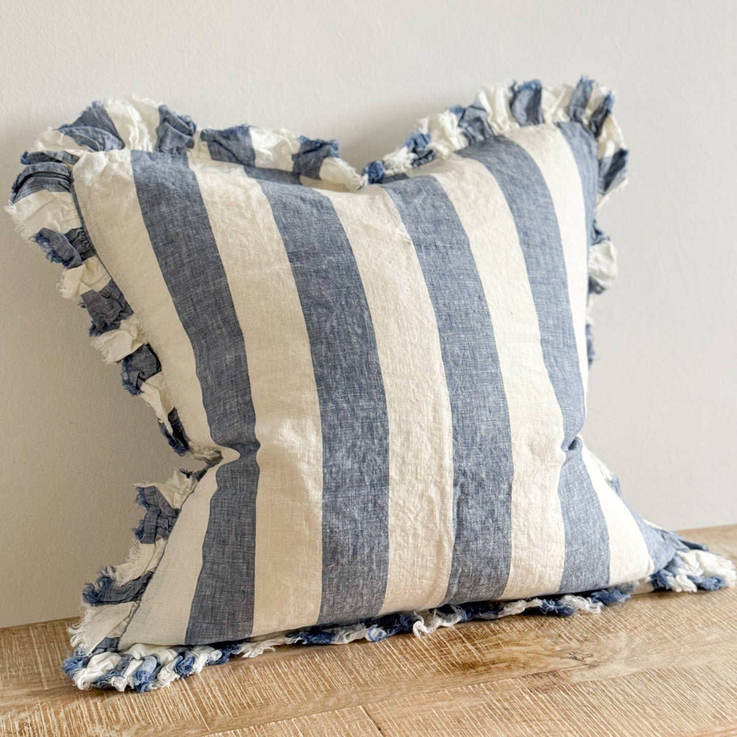 Ellenvale Hallie Ruffled Edged Linen Cushion in Blue Wide Stripe displayed on a bench against a white wall.