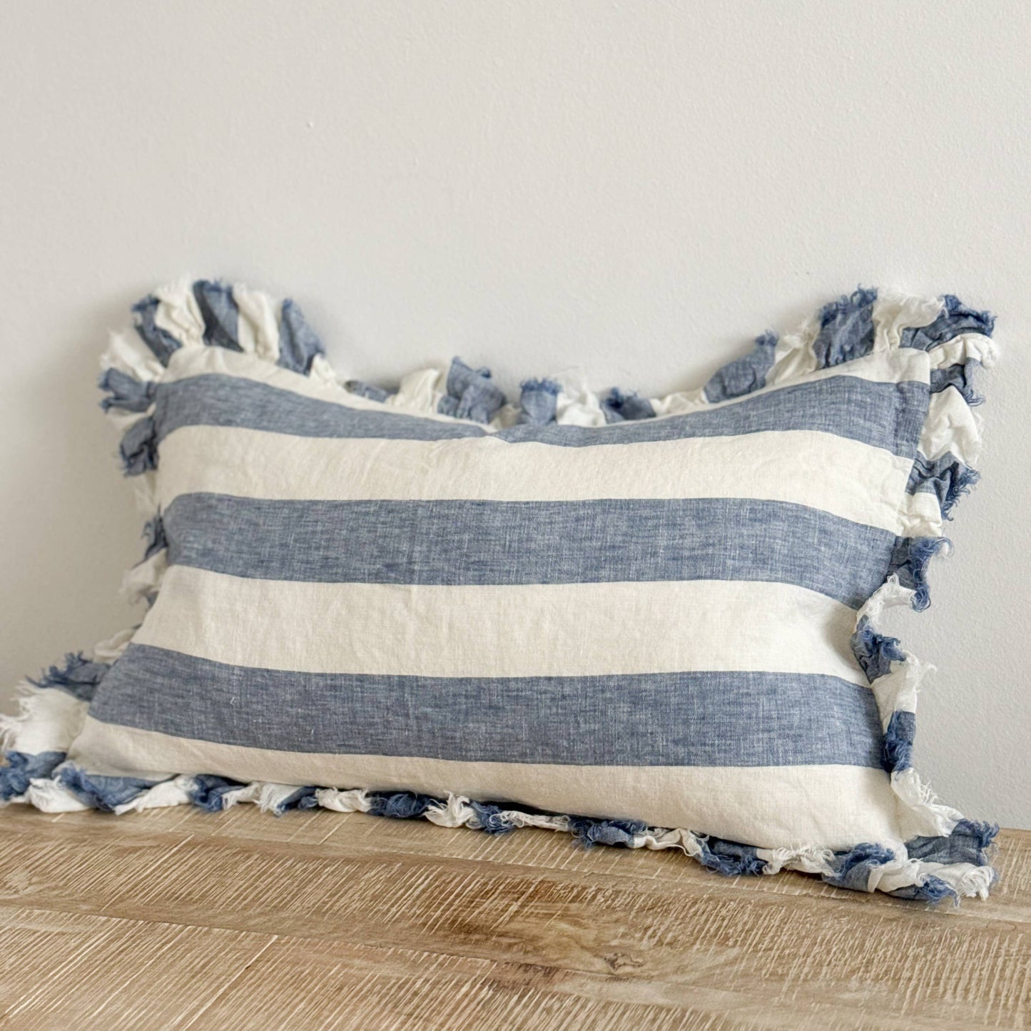 Ellenvale Hallie Ruffled Edged Linen Cushion in Blue Wide Stripe displayed on a bench against a white wall.
