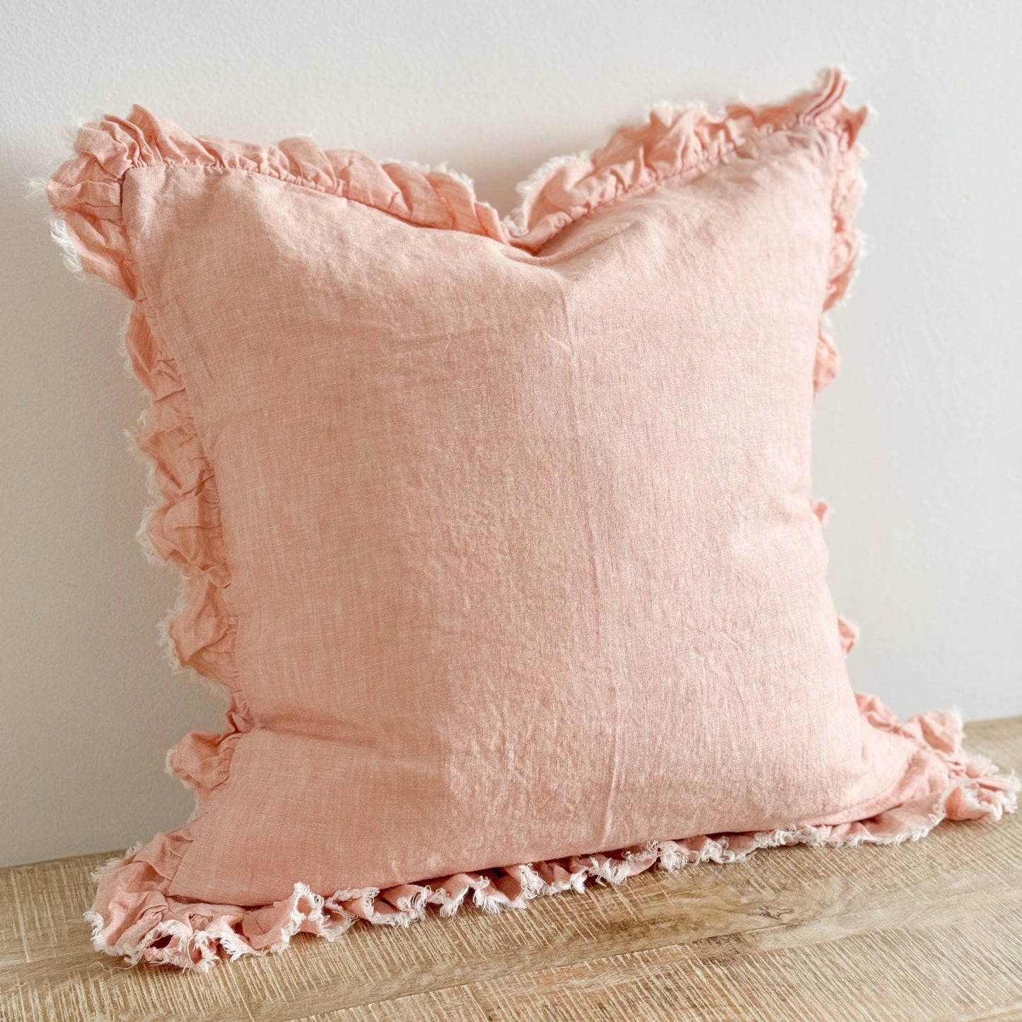 Ellenvale Ruffled Edge Linen Cushion in Washed Pink displayed on a bench against a white wall.