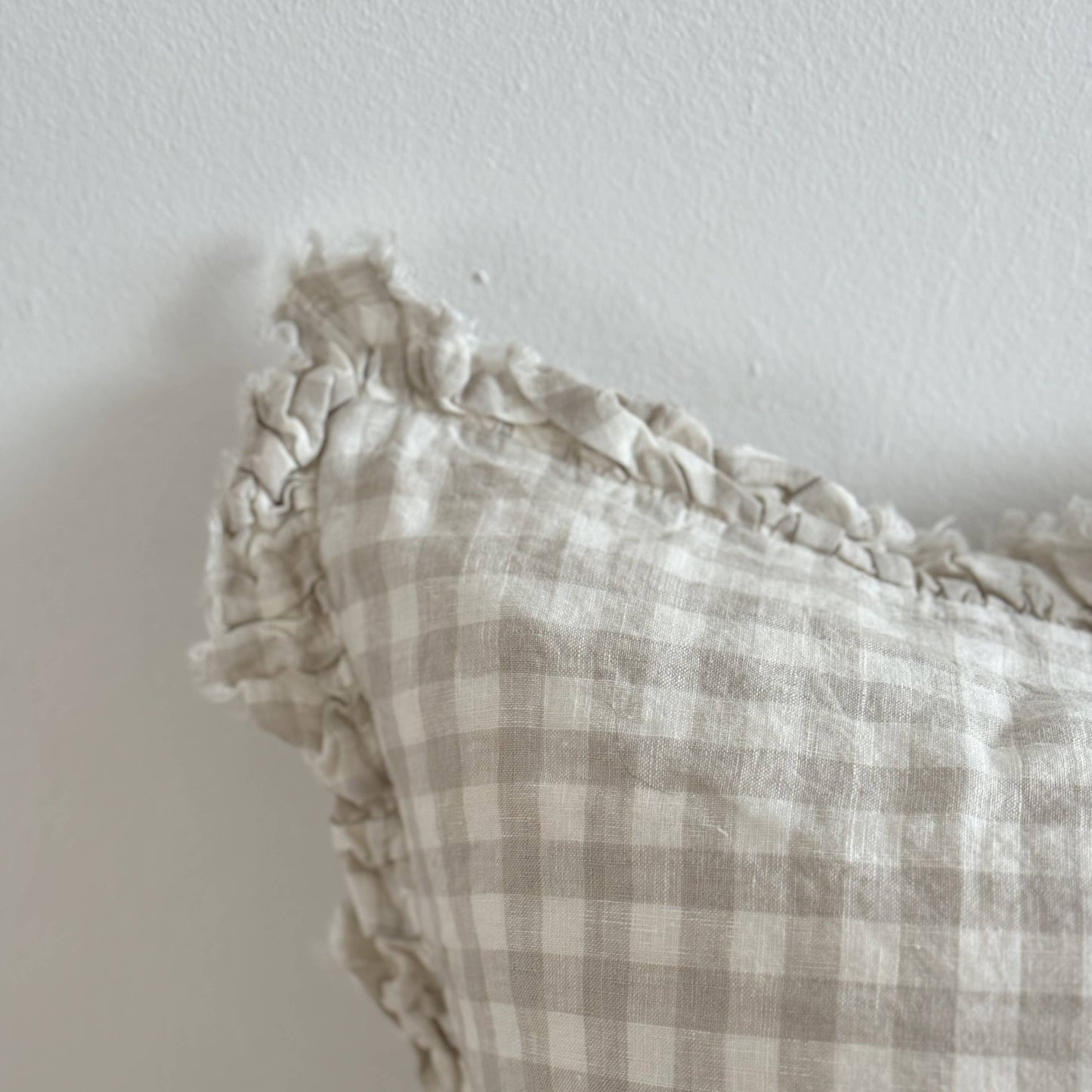 Ellenvale Ruffled Edged Linen Cushion Cover in Natural Gingham displaying the corner edge of the cushion against a white wall.