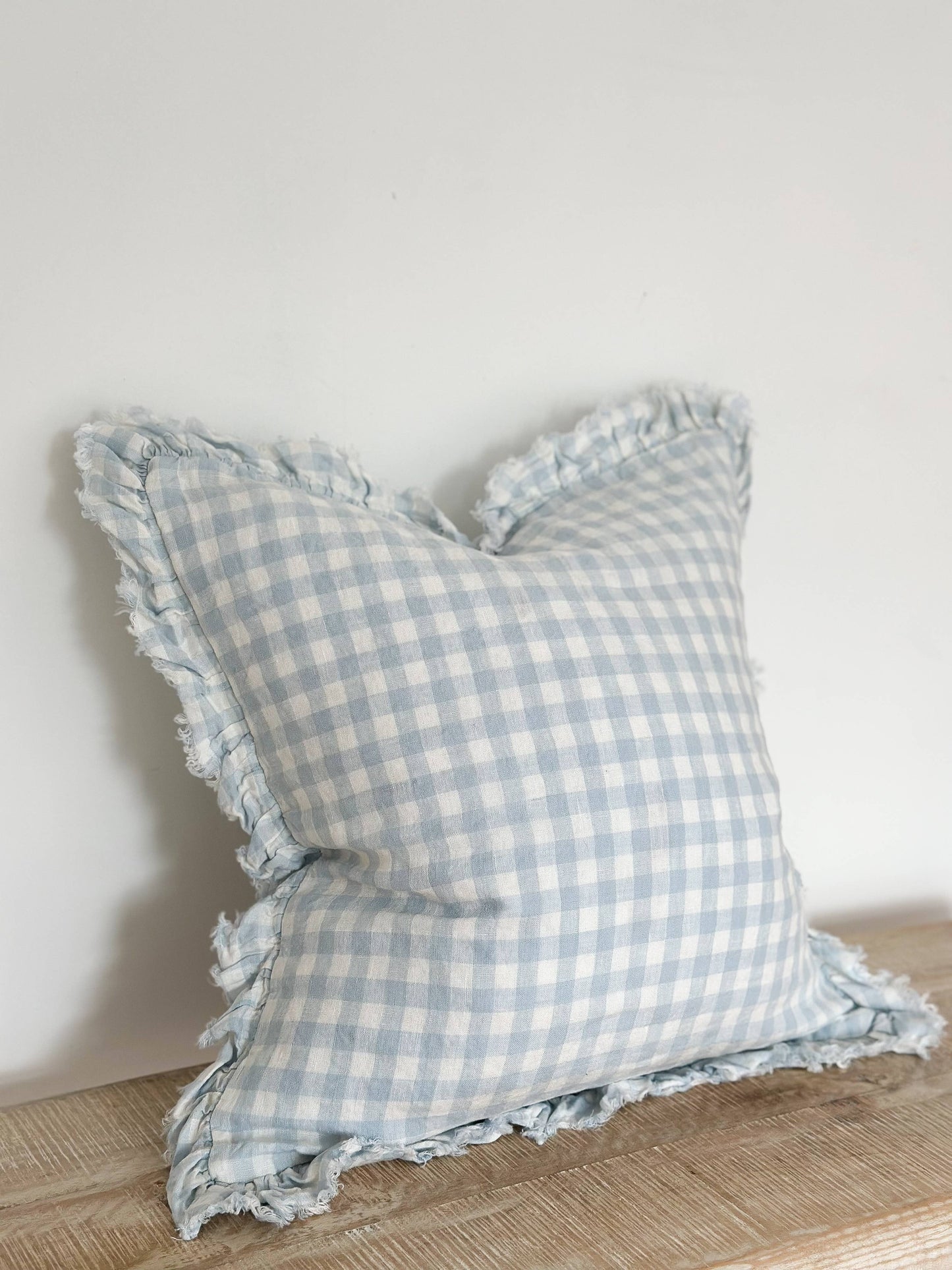Ellenvale Ruffled Linen Cushion in Blue Gingham with a ruffled edge displayed on a bench against a white wall.