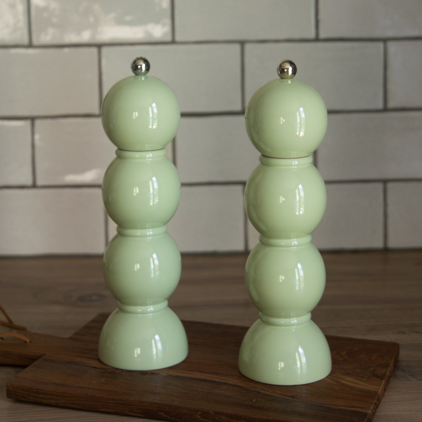 Two light green salt and pepper shakers on a wooden board with a tiled wall background.