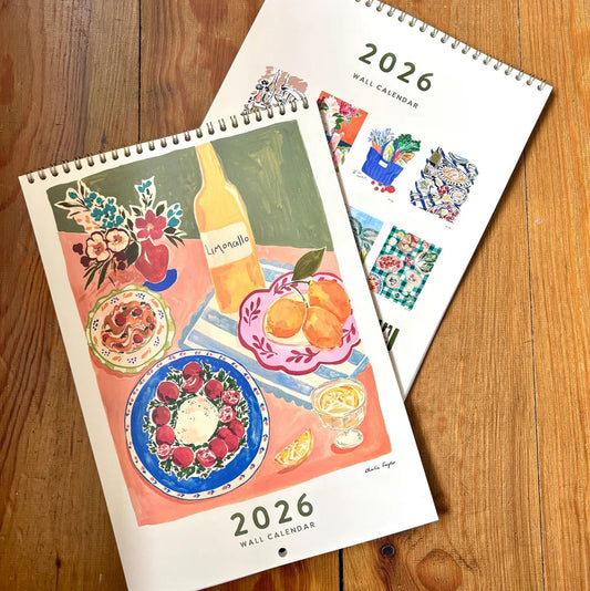 Two 2026 wall calendars on a wooden surface, one with a colorful illustration of food and drink, the other with a floral design.