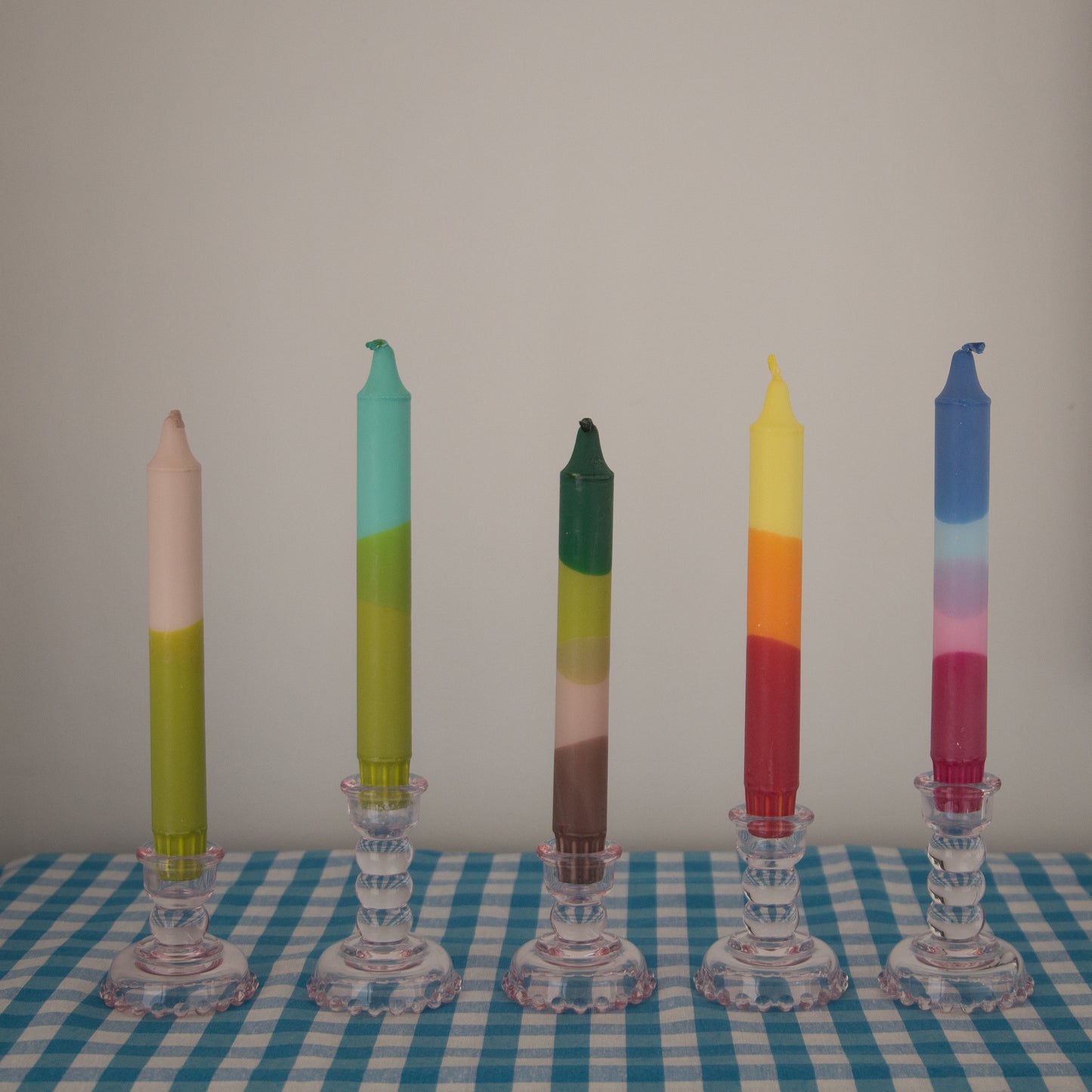 Nordtrice Ombre Dipped Tapered Candles in different colours displayed with other candles in pink glass candle holders on a blue gingham cloth