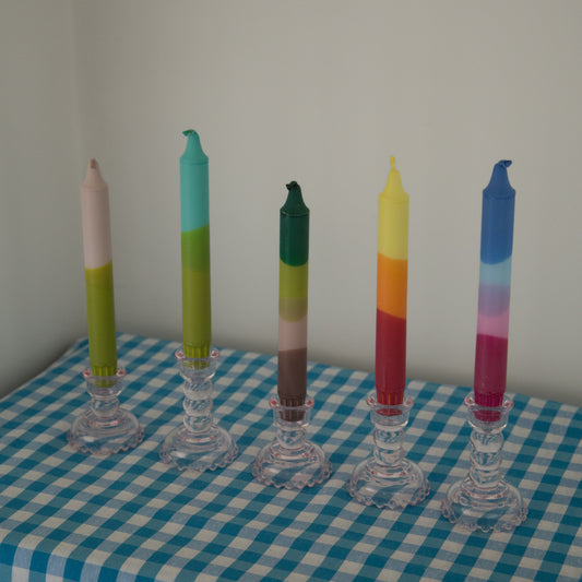 Five handmade ombre dipped taper candles in clear glass holders line a blue and white checkered cloth against a gray backdrop. Keywords: ombre, dipped, taper candles, handmade, colorful, glass holders, checkered cloth.