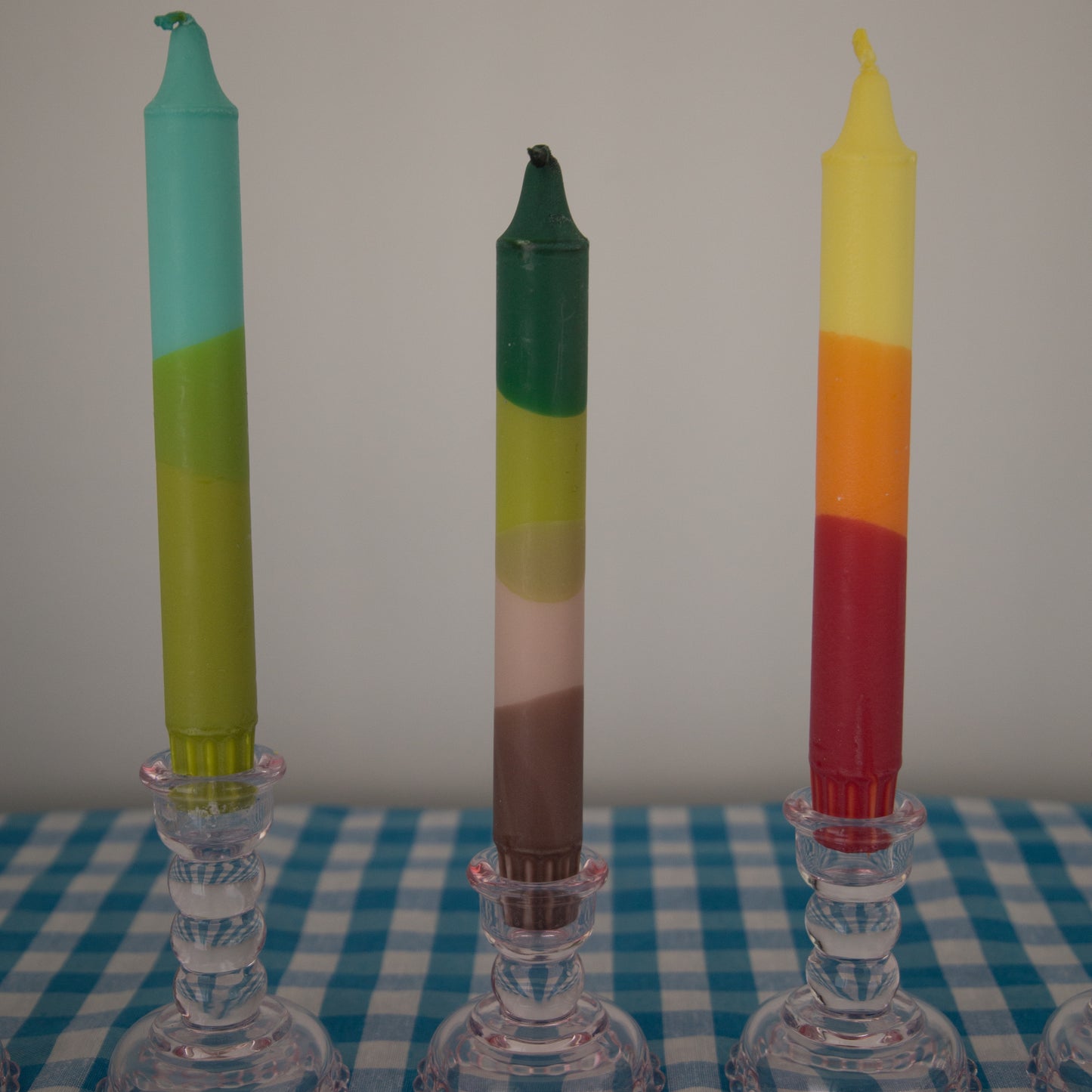 Nordtrice tapered candles in multi colours dipped and ombre displayed in a pink glass candle holder on a blue gingham cloth