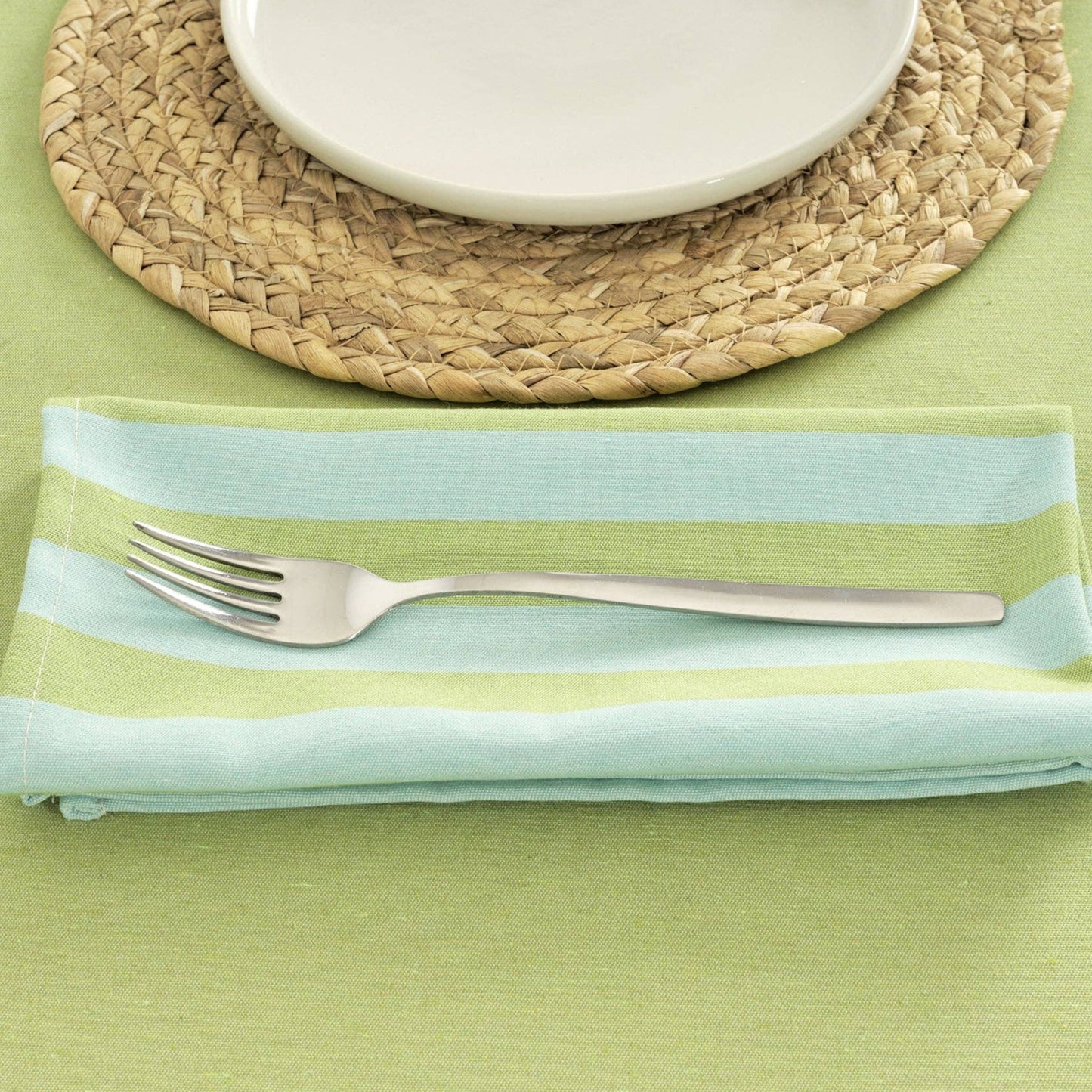 Pistachio and Turquoise striped table napkin, folded next to a woven placemat and white plate. The napkin is displaying a fork on a green tablecloth.
