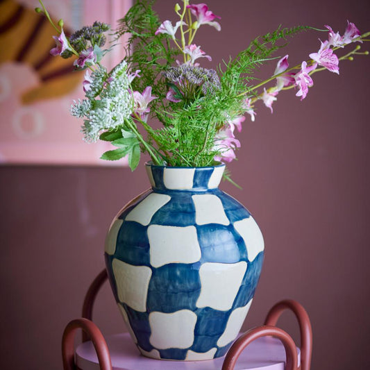 Blue and white checkered vase with flowers on a pink stool against a pink wall