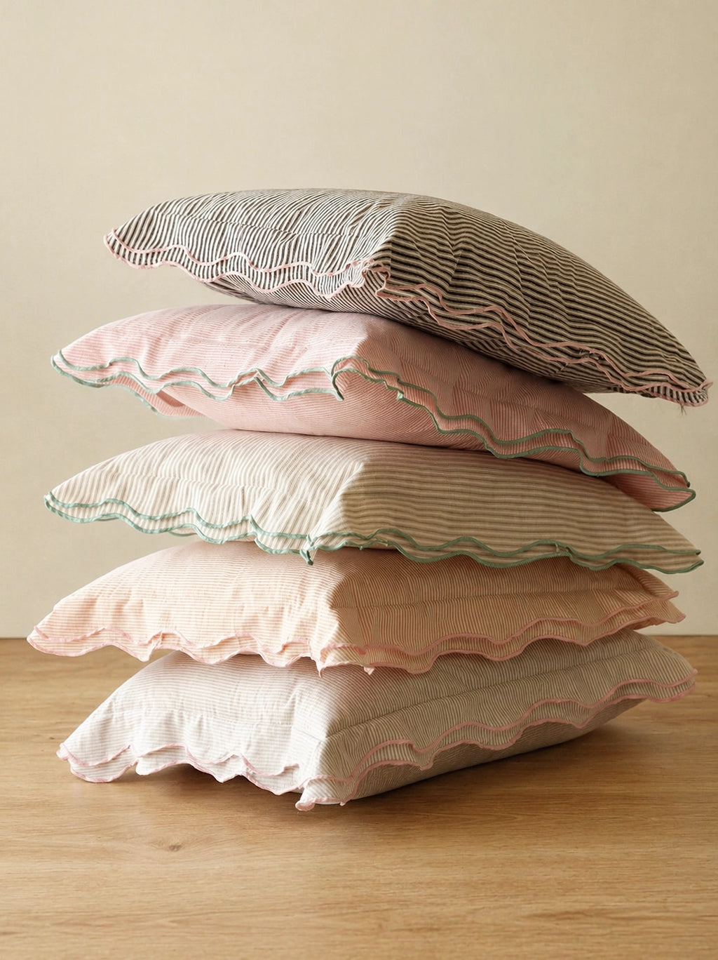 A stack of five decorative throw pillows in soft pastel colors, including pink, beige, and gray, with striped and solid fabric textures and scalloped edges, displayed on a wooden surface against a neutral beige background.