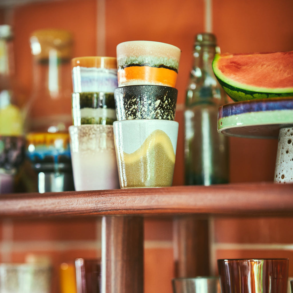 Stacked glasses with colorful bases on a wooden shelf against an orange wall.