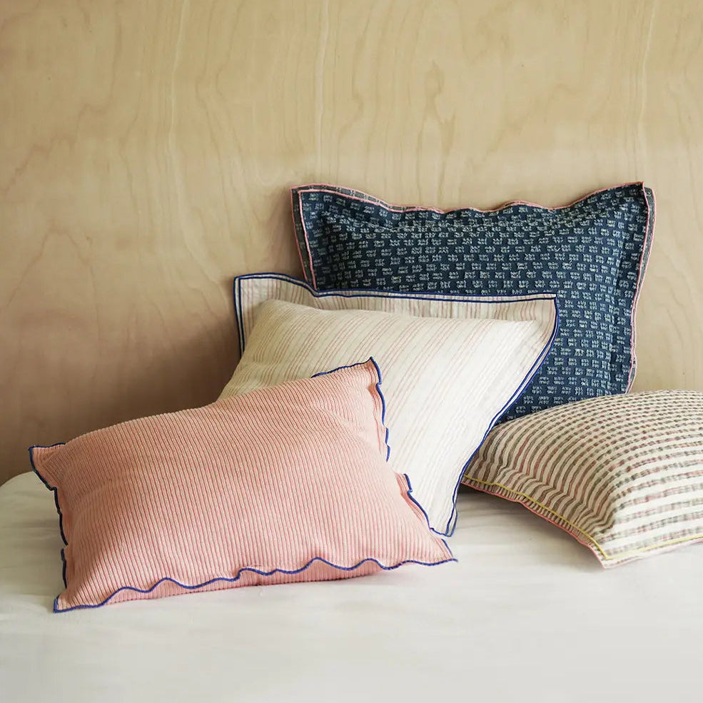 Four decorative throw pillows arranged on a bed against a light wood headboard. The pillows include a dusty pink striped pillow with blue piping in front, a cream striped pillow behind it, a dark blue patterned pillow at the back, and a beige striped pillow on the right.