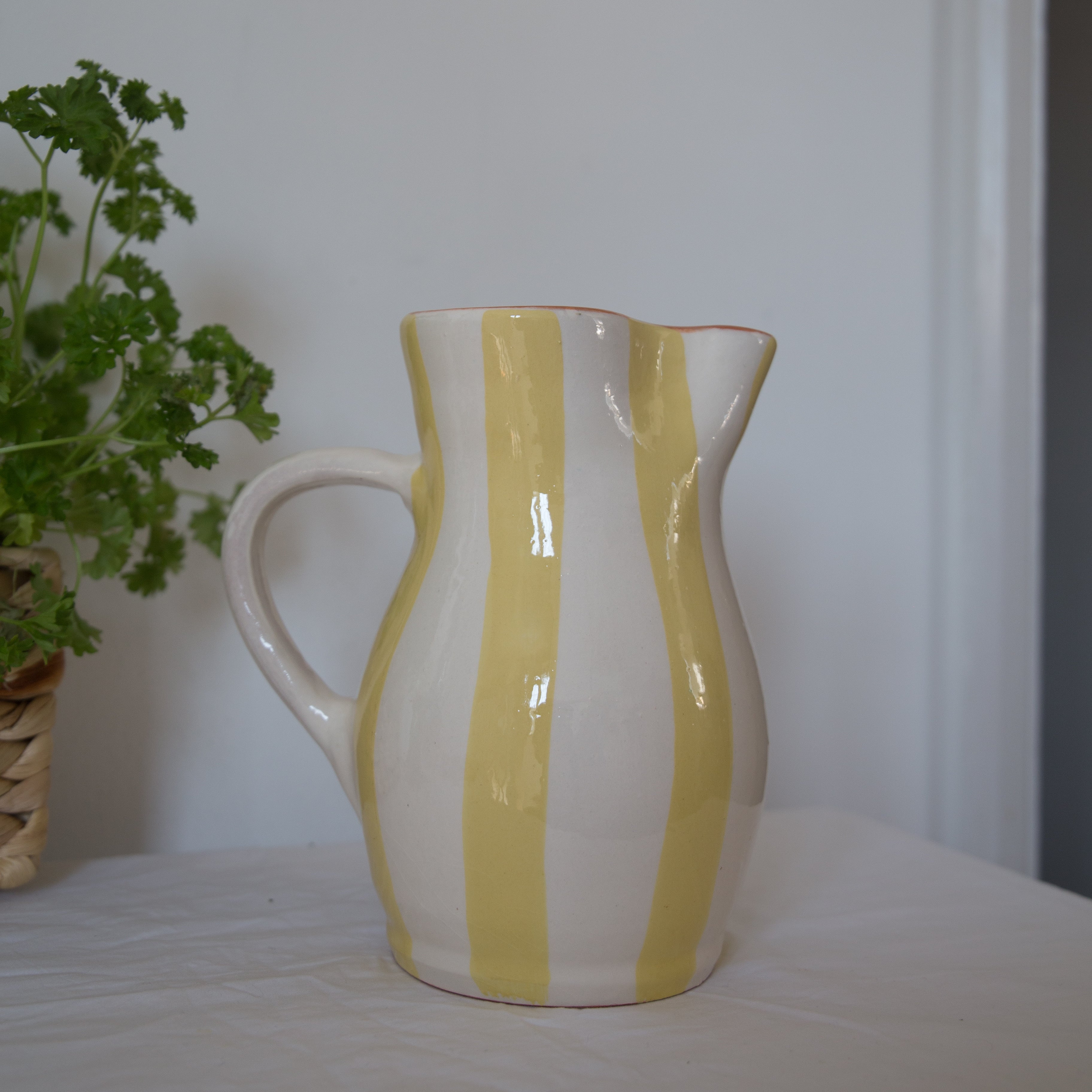 A handcrafted ceramic jug with yellow stripes on a white background, with a terracotta interior, displayed on a table with a plant decoration in the background.