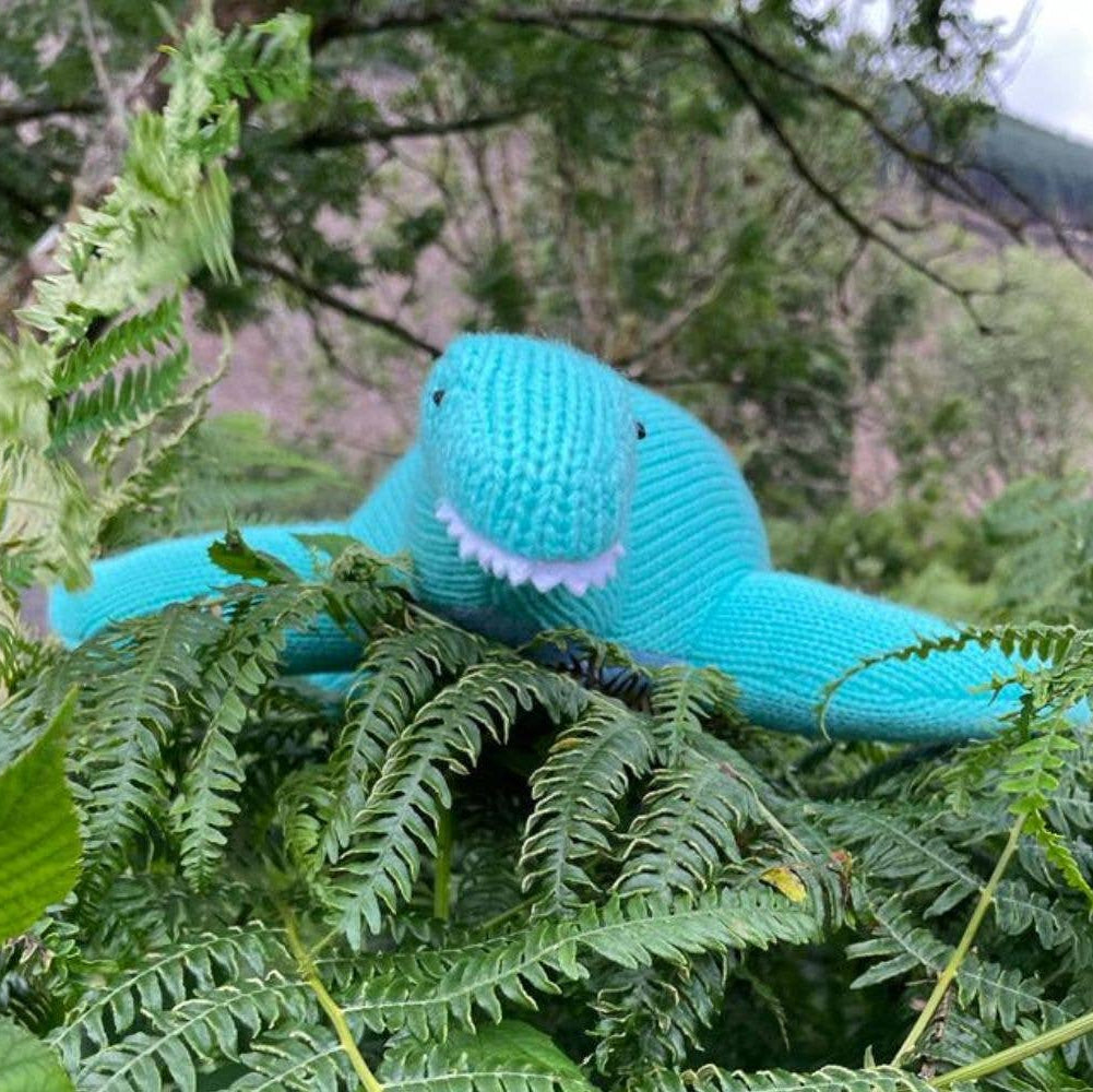 Knitted blue dinosaur toy among green ferns with a blurred natural background