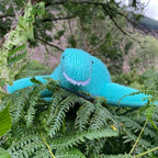 Knitted blue dinosaur toy among green ferns with a blurred natural background