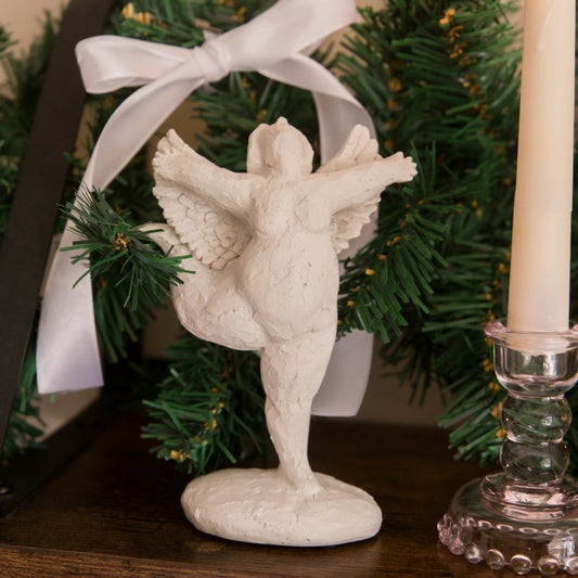 White angel figurine with outstretched wings on a dark surface with greenery and a candle in the background.