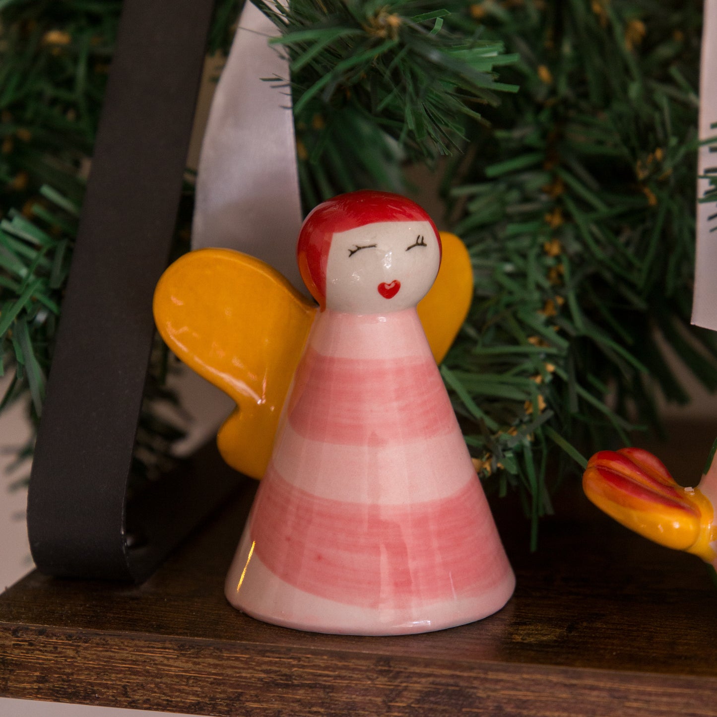 Ceramic angel figurine with red hair and yellow wings on a wooden surface with a blurred Christmas tree background.