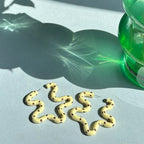 Gold snake-shaped earrings on a light surface with a green vase and plant in the background.