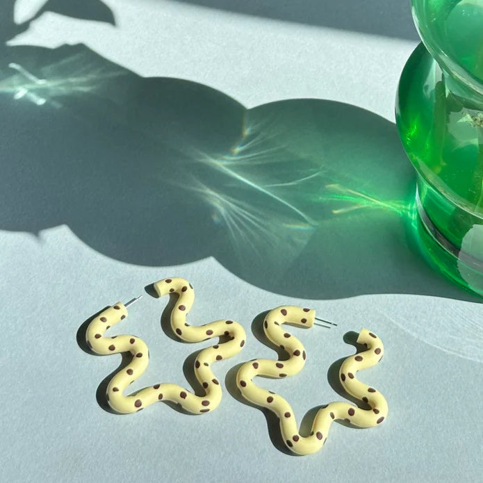 Gold snake-shaped earrings on a light surface with a green vase and plant in the background.