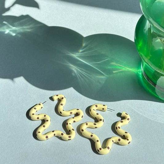 Gold snake-shaped earrings on a light surface with a green vase and plant in the background.