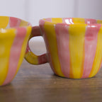Two Bloomingville nini Cups in Pink and Yellow Stripe displayed next to each other on a bench with a white background.
