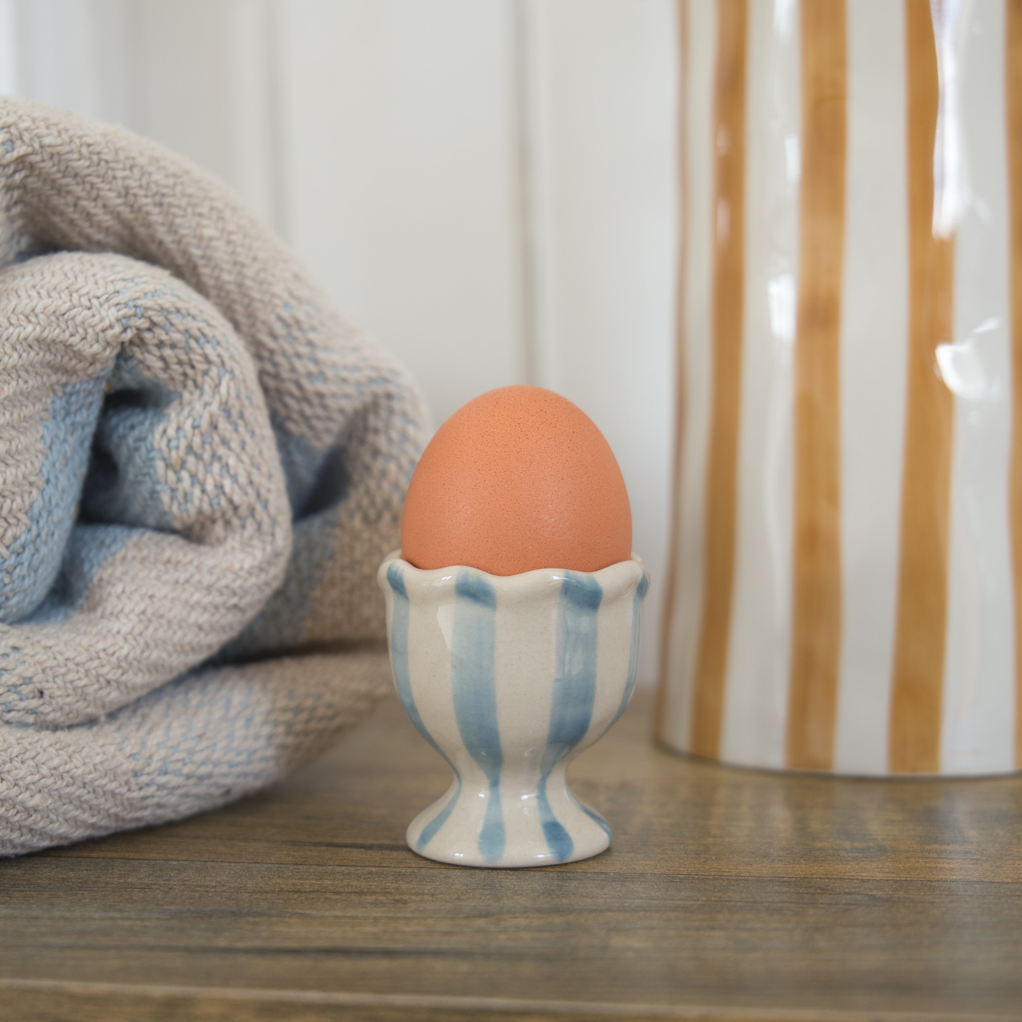 Bloomingville Willa Egg Cup in blue and white stripe is displaying an egg on a table with a blue and white striped throw accompanied with a brown and white striped ceramic jug in the background.