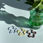 Three unique earrings on a light surface with a green vase and flowers in the background.
