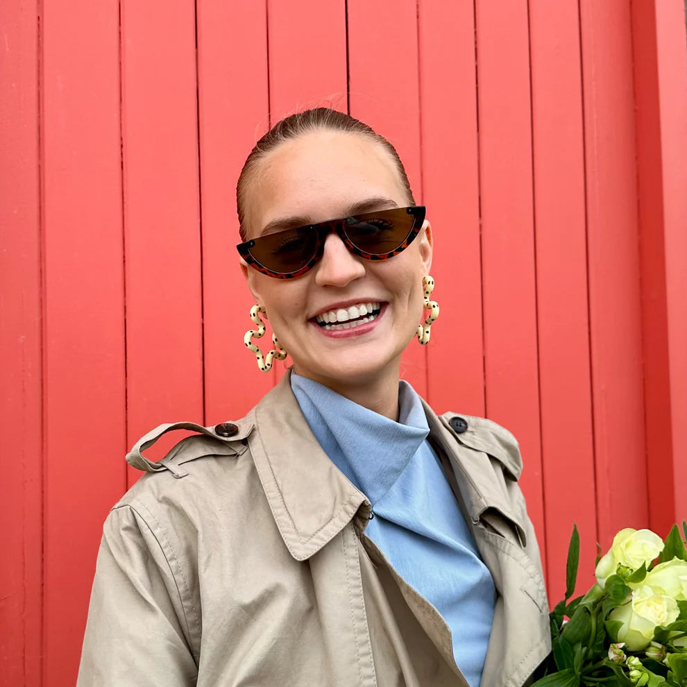 Woman in a beige trench coat and sunglasses holding flowers against a red background