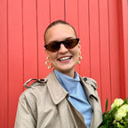 Woman in a beige trench coat and sunglasses holding flowers against a red background