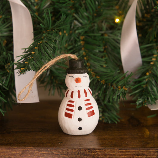 Small snowman ornament with a black top hat and red scarf on a wooden surface with a Christmas tree in the background.