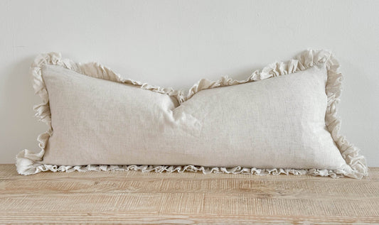 Ellenvale Grace Linen Blend Cushion Cover in Sand colour.  This is a bolster cushion with a ruffled edge displayed on a bench against a white wall.  
