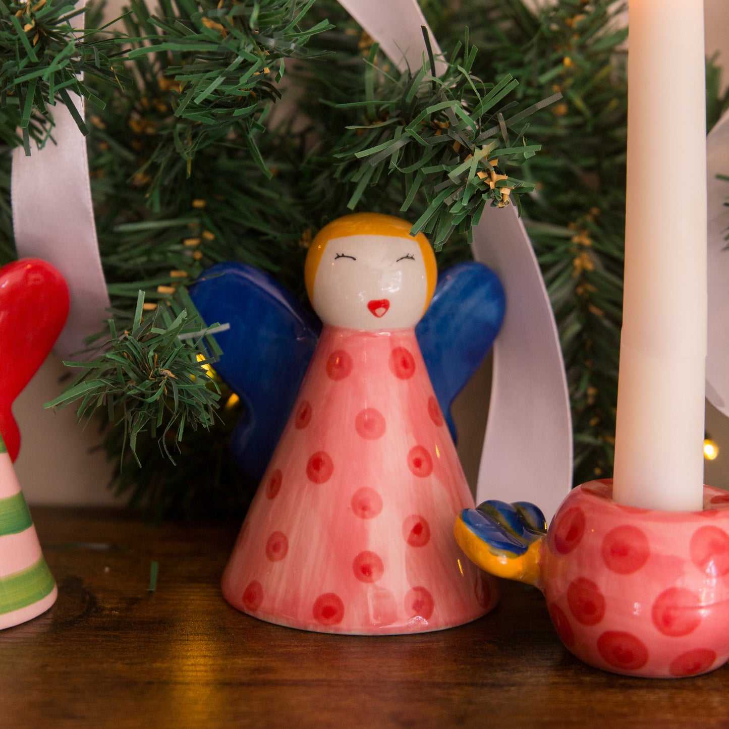 Decorative angel figurine with red polka dots and a candle holder on a wooden surface with a Christmas tree in the background.