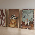 Three framed artworks on a wooden floor against a plain wall.