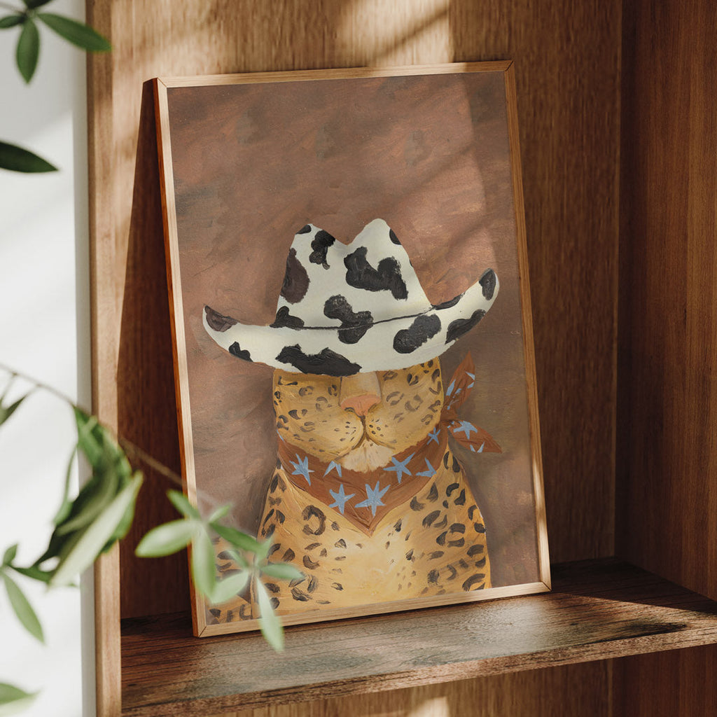 Framed artwork of a cat wearing a cowgirl hat on a wooden shelf.