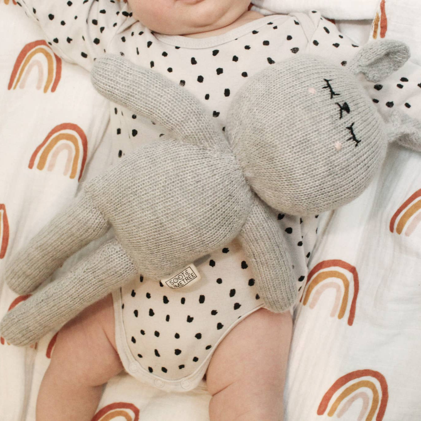 Lenny the Handmade Grey Cuddle Bunny laying on a baby with a rainbow muslin in the background.