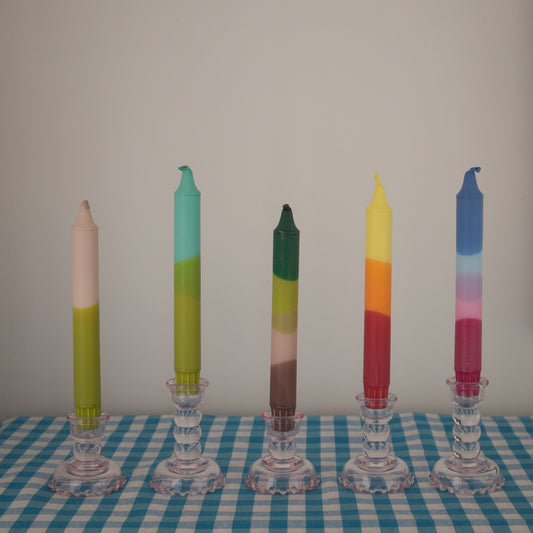 Nordtrice Ombre Dipped Tapered Candles in different colours displayed with other candles in pink glass candle holders on a blue gingham cloth
