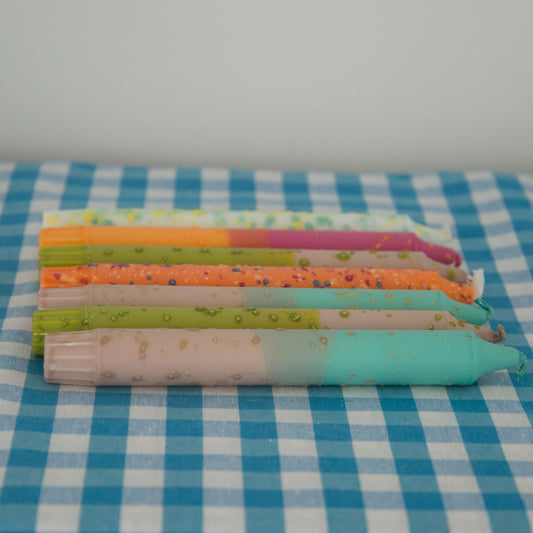 Nordtrice confetti and ombre tapered candles lying down in a row on a gingham table cloth 