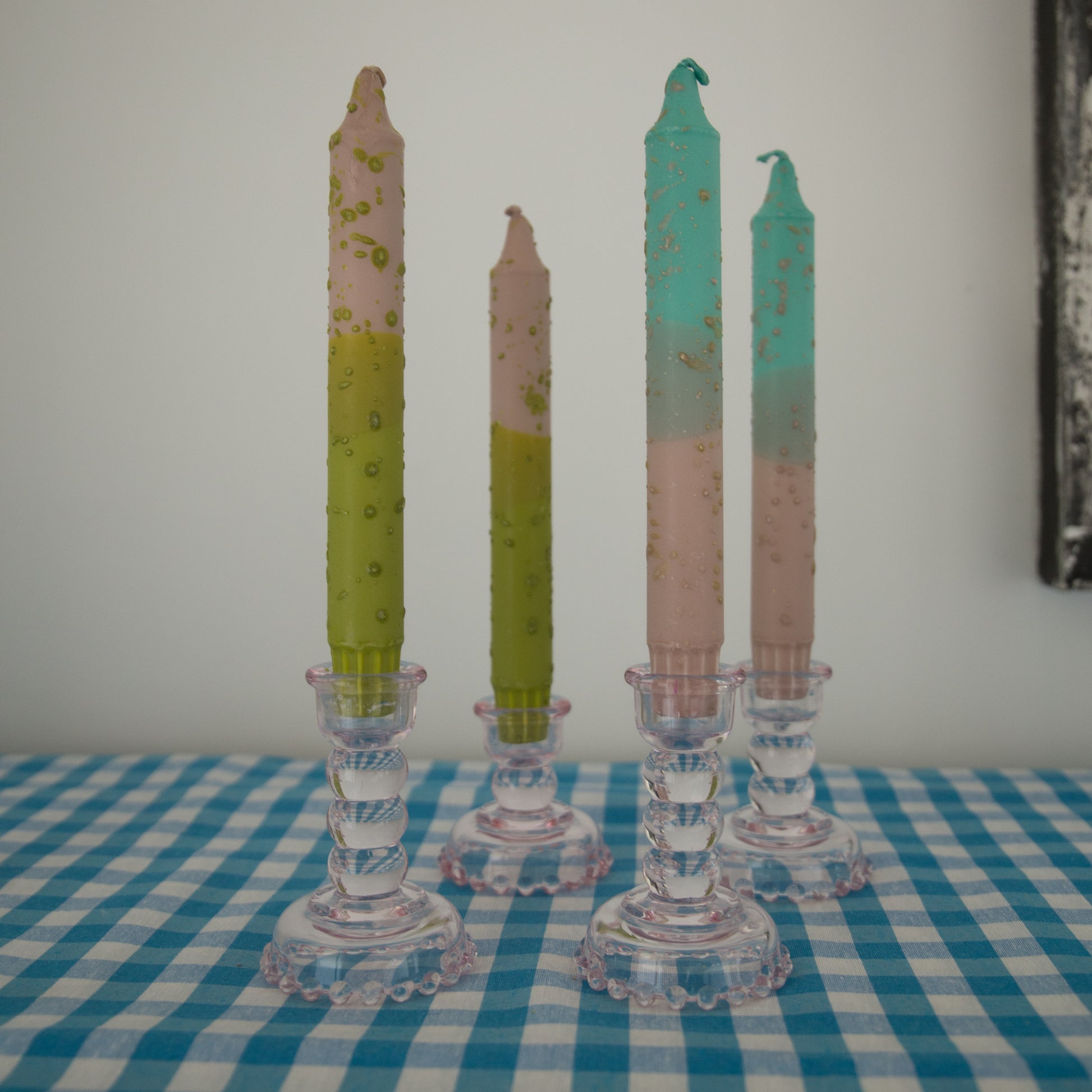 Nordtrice confetti tapers in greens and pinks displayed in a pink glass candle holder on a blue gingham cloth 