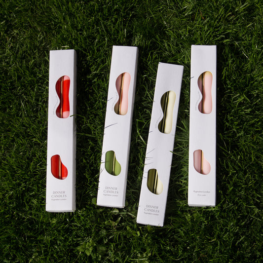 set of two dinner candles displayed in their boxes lay next to each other on the grass. Showing the front packaging and the styles in stock. 