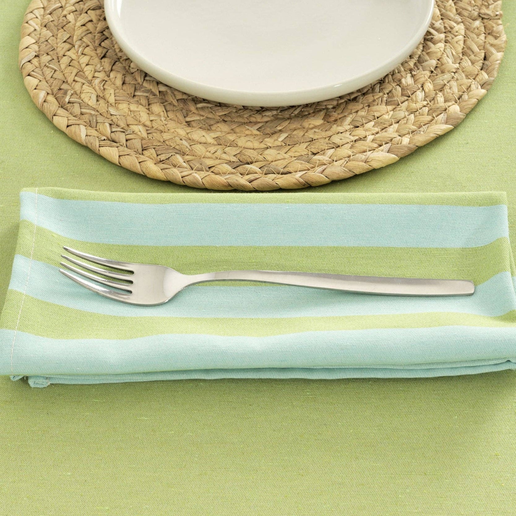 Pistachio and Turquoise striped table napkin, folded next to a woven placemat and white plate.  The napkin is displaying a fork on a green tablecloth.