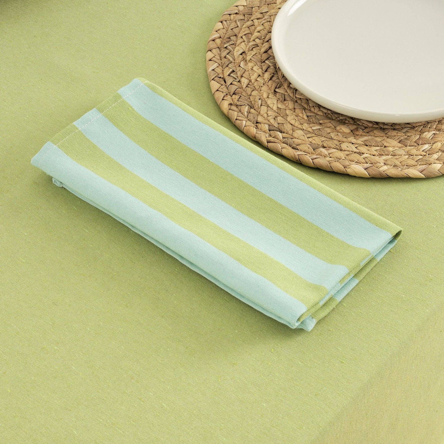 Table napkins in pistachio and turquoise laid on a green tablecloth, next to a woven placemat and white plate.
