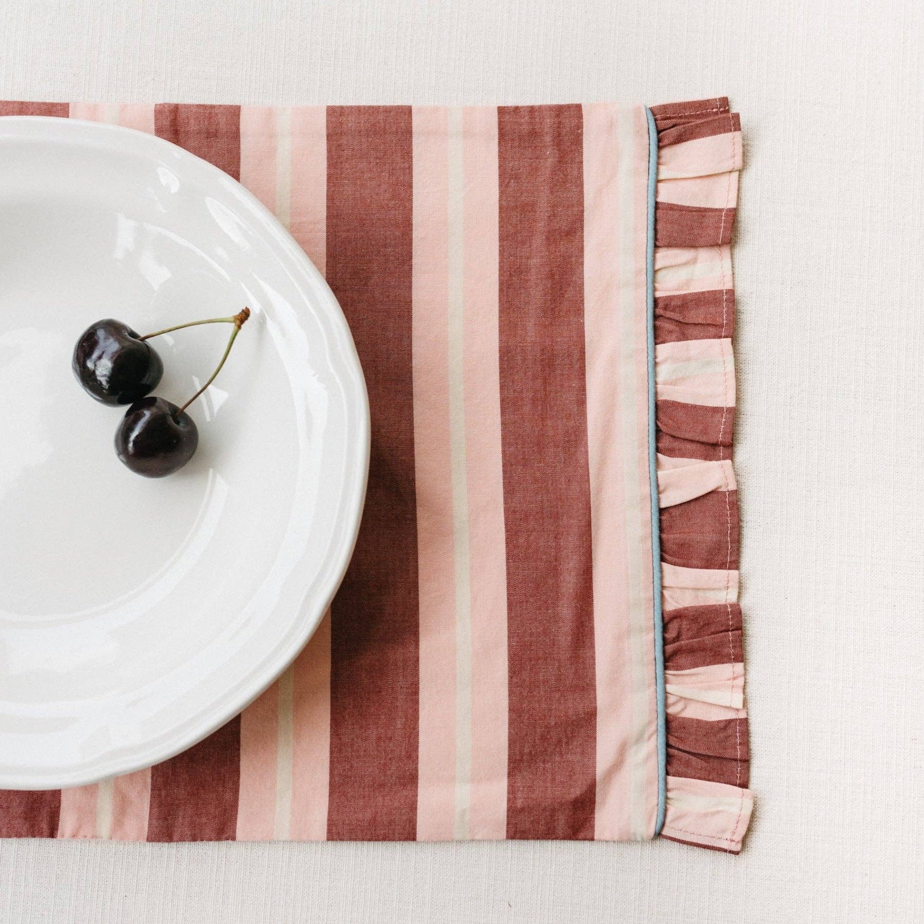 Pink and Red stripe cotton placemat displayed with a white plate with 2 cherries placed on top.
