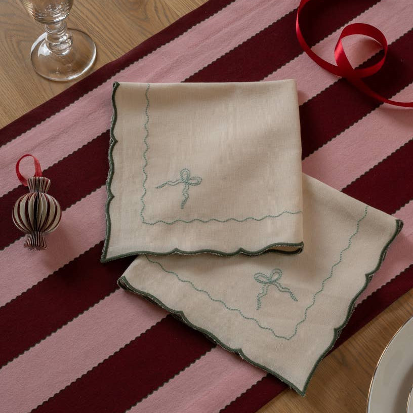 Beige embroidered napkins on a striped pink and red table runner with a glass and decorative item.