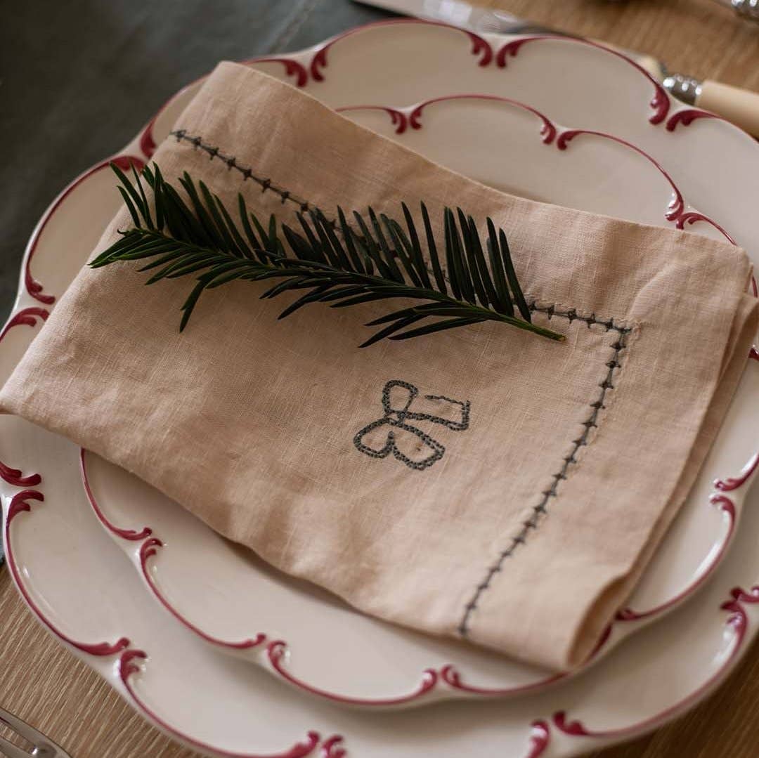Folded beige napkin with embroidered design on a set of white plates with red trim.