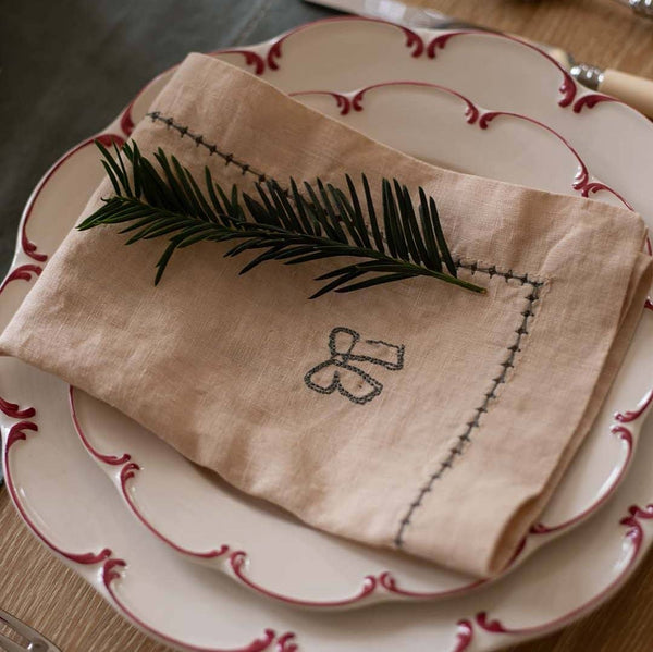 Folded beige napkin with embroidered design on a set of white plates with red trim.