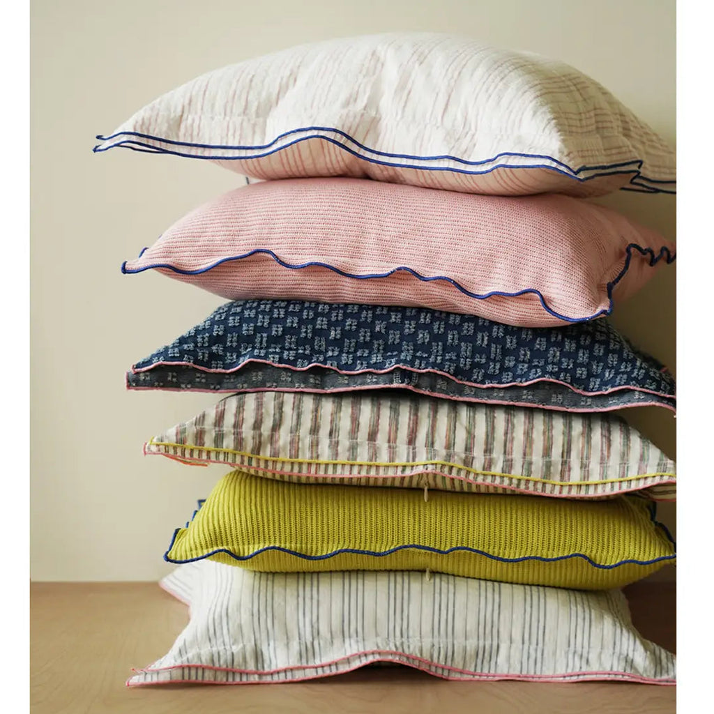 A stack of five decorative pillows in different colors and patterns, including cream, pink, navy, beige, and yellow, with scalloped edges, set against a neutral beige background.