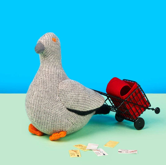 Knitted pigeon pulling a small red cart with items on a blue and green gradient background